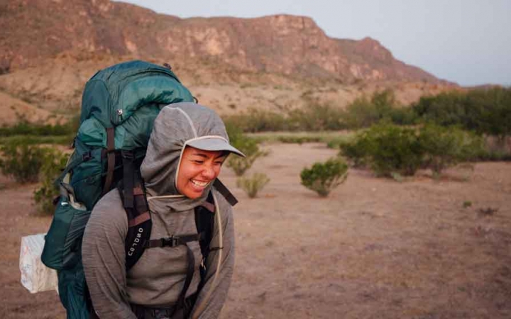 a woman wearing a backpack laughs in front of a mountainous desert landscape